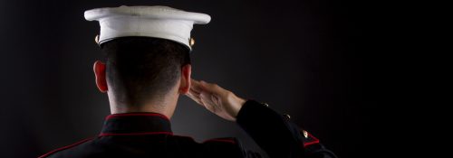 A U.S. Marine saluting against a black background while in the dress blues service uniform.