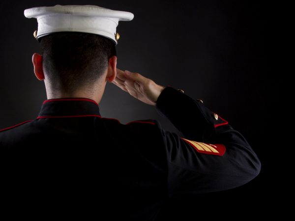 A U.S. Marine saluting against a black background while in the dress blues service uniform.
