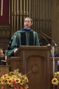 Adam Carrington in doctoral robes giving a speech at commencement.