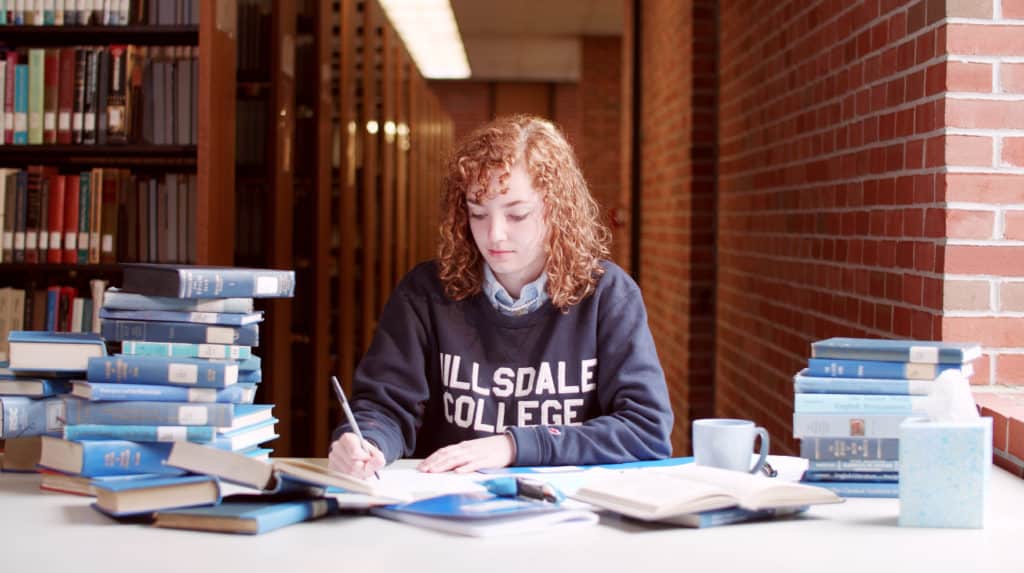 Hillsdale College student writing in the library, surrounded by blue books.