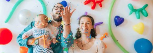Parents celebrate in front of a bright, colorful background of shaped balloons