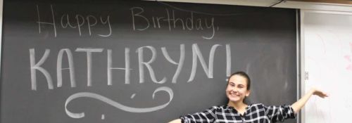 Female student stands smiling in front of a blackboard that reads 