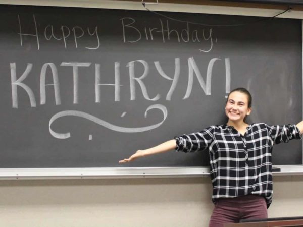 Female student stands smiling in front of a blackboard that reads "Happy Birthday, Kathryn!"