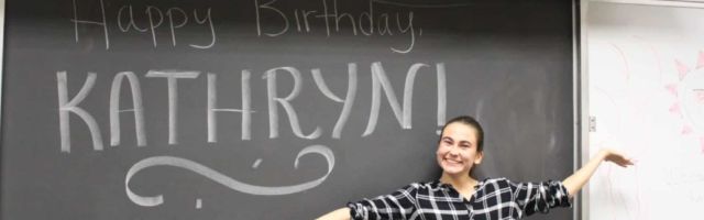 Female student stands smiling in front of a blackboard that reads "Happy Birthday, Kathryn!"