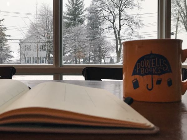 A book and a mug sitting on a table in front of a window overlooking a snowy day